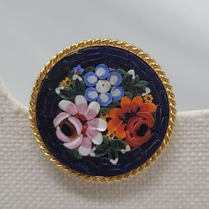May include: Round mosaic brooch featuring a floral design with pink, white, orange, and blue flowers on a dark blue background. The brooch has a gold-colored rope-like border. A close-up shot highlights the intricate details of the mosaic.