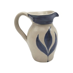 May include: A ceramic pitcher with a beige body and a dark blue rim and floral design. The pitcher has a curved handle and a spout for pouring. The floral design is a stylized depiction of leaves.