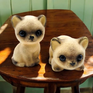 May include: Two Siamese cat figurines with large, dark blue eyes and tan fur. One cat is sitting upright, the other is lying down. The figurines have black accents on their ears, faces, and paws. They are displayed on a wooden chair.