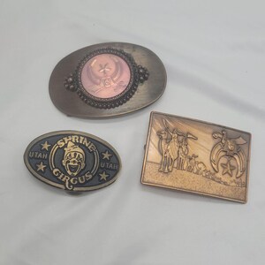 May include: Three vintage belt buckles. One oval buckle with a pink center and decorative border. Another oval buckle with "Shrine Circus Utah" text and a clown face. The third is a rectangular buckle with a camel caravan.