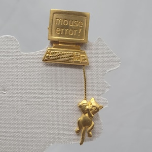May include: A gold-colored computer-themed pin. The pin features a computer monitor with the words "mouse error!" and a keyboard. A gold-colored cat is hanging from the keyboard.