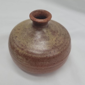 May include: A small, round, ceramic vase with a narrow neck. The vase has a speckled, earthy color palette of brown, tan, and rust. The surface has a textured appearance, with visible rings around the body.