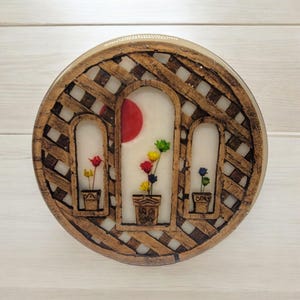May include: A round, decorative piece featuring a lattice design in shades of brown. Three arched windows display colorful flowers in pots, with a red sun in the background. The piece is likely a wall hanging or decorative item.