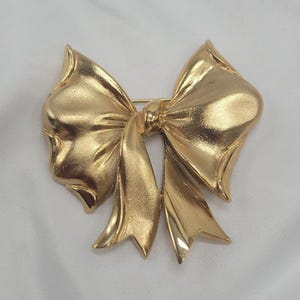 May include: A gold-colored bow brooch with a pin closure. The brooch is made of a shiny, metallic material and features a detailed bow design with textured folds and curves. The bow is a classic accessory.