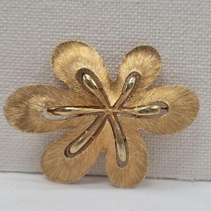 May include: A gold-tone flower brooch with six textured petals and a detailed center. The brooch has a brushed finish, giving it a vintage look. The petals are slightly curved, adding dimension to the design. A classic accessory.