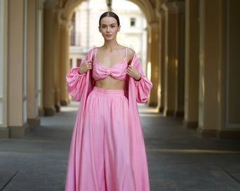May include: A three-piece pink outfit consisting of a crop top, wide-leg pants, and a long-sleeved robe. The crop top has a bow detail at the bust. The outfit is made of a flowing, silky fabric. The model is standing in an arched walkway.
