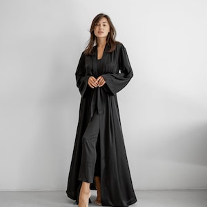 May include: A black satin robe with wide sleeves and a matching belt. The robe is paired with black trousers and a top. The outfit is worn by a person standing in front of a white wall.