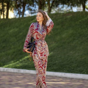 May include: A burgundy paisley print pantsuit with a matching wrap top and wide-leg pants. The outfit features a belt, gold jewelry, and a black handbag with a gold chain. The ensemble is completed with red shoes.