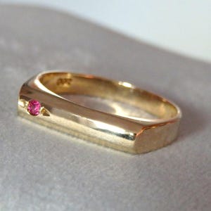Peut inclure: Une bague en or avec un anneau rectangulaire et une petite pierre précieuse rose ronde incrustée sur le dessus. La bague a une finition polie, reflétant la lumière. La pierre précieuse est d'un rose vif, contrastant avec l'or.