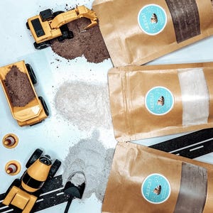 Pode incluir: Uma disposição plana com veículos de construção de brinquedo, incluindo um camião basculante, uma escavadora e uma betoneira, juntamente com pequenos cones de trânsito laranja. Vários sacos de papel castanho com etiquetas circulares azuis e brancas também estão presentes, com vários pós coloridos espalhados.