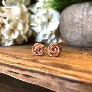 May include: A pair of rose-gold colored wire earrings shaped like roses. The earrings are on a wooden surface.
