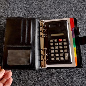 May include: Open black leather binder with a black calculator inside. The calculator has white buttons and a digital display. The binder also contains colorful divider tabs and a clear pocket.