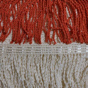 Peut inclure: Gros plan sur une frange décorative aux tons orange et crème. La frange orange présente un motif de corde torsadée, tandis que la frange crème est de style similaire. La frange est fixée à un support tissé, adapté à divers projets d'artisanat.