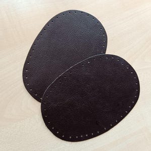 Könnte beinhalten: Zwei braune Lederflicken mit einem genähten Rand. Die Flicken sind oval geformt und haben eine strukturierte Oberfläche.