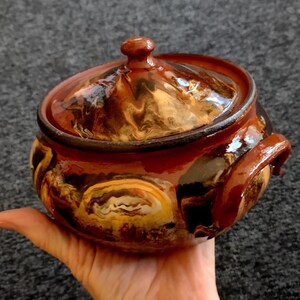 Puede incluir: Una olla de cerámica hecha a mano con tapa. La olla es principalmente marrón con remolinos de amarillo y negro, y un acabado brillante. Tiene un pequeño pomo en la tapa y un asa en el lateral.