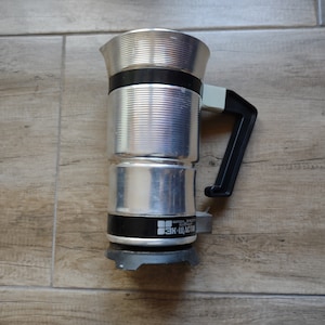 May include: A silver and black stovetop espresso maker with a black handle. The maker has a label that reads "3K-ЩАСТЕ" in Cyrillic letters.