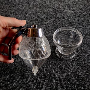 Könnte beinhalten: Ein klarer Honigspender aus Glas mit Chromgriff und Deckel, der ein Wabenmuster aufweist. Daneben steht eine kleine, klare Glasschale. Der Spender ist für einfaches Eingießen von Honig konzipiert.