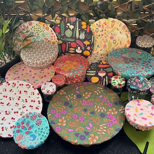 Op de afbeelding: Een verzameling herbruikbare voedselhoezen met diverse patronen, waaronder kersen, groenten en bloemen. De ronde hoezen zijn gemaakt van stof en verkrijgbaar in verschillende maten, geschikt voor het afdekken van kommen en schalen.