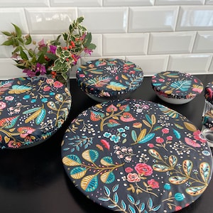Puede incluir: Un juego de seis cubiertas de alimentos reutilizables con un patrón floral sobre un fondo negro. Las cubiertas están hechas de tela y tienen un cierre de cordón. Se muestran sobre una encimera de azulejos blancos.