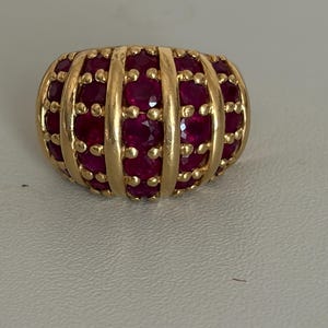 Op de afbeelding: Een goudkleurige ring met een afgeronde vorm, met verticale gouden banden en rijen kleine, levendige rode edelstenen. Het ontwerp van de ring is symmetrisch, met de edelstenen gelijkmatig verdeeld.
