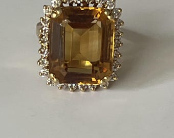 Pode incluir: Um anel de ouro com uma grande pedra preciosa retangular de cor âmbar. A pedra preciosa é rodeada por um halo de pedras preciosas menores e transparentes. O anel está sobre um fundo branco.