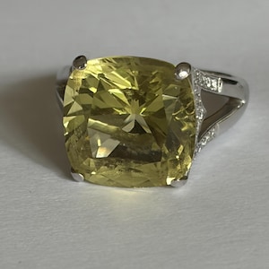 May include: A ring featuring a large, cushion-cut, pale yellow gemstone held by four silver-toned prongs. The band is made of a polished silver-toned metal and includes a split-shank design accented with small, clear, round-cut stones along the sides.