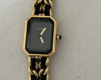 Könnte beinhalten: Eine goldfarbene Armbanduhr mit einem schwarzen rechteckigen Zifferblatt und einem schwarzen Leder- und goldfarbenen Kettengliederarmband. Das Zifferblatt hat goldene Zeiger und das Wort "SWISS" ist unten aufgedruckt. Der Markenname "CHANEL" ist oben aufgedruckt.