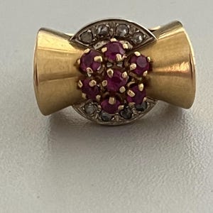 May include: A gold-toned ring with a central cluster of small, round, red gemstones. The ring features a bow-like design with a band of small, clear gemstones above the red cluster. The ring's design is symmetrical and elegant.