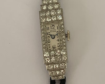 Könnte beinhalten: Eine Vintage-Armbanduhr mit einem diamantbesetzten Gehäuse und einem schwarzen Lederarmband. Das Zifferblatt der Uhr ist weiß mit schwarzen römischen Ziffern für die Stunden und dem Text "ACETO SUISSE" in der Mitte.