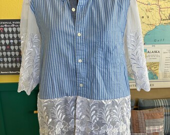 May include: A button-down shirt with blue and white vertical stripes, featuring sheer white lace sleeves and a scalloped lace hem. The shirt is displayed on a mannequin, showcasing a unique design with a blend of textures and patterns.