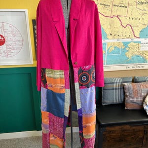 May include: A long, open-front coat in a vibrant fuchsia color. The coat features a patchwork design in shades of orange, blue, purple, and pink. The coat is displayed on a mannequin, showcasing its tailored silhouette and unique textile combination.