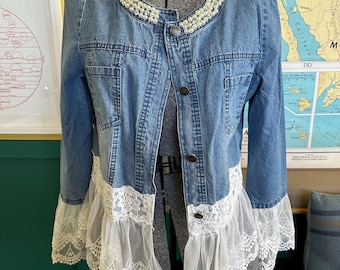 May include: A blue denim jacket featuring a round neckline embellished with white faux pearls. The jacket has a button-front closure and long sleeves. The hem and sleeve cuffs are finished with a wide, white sheer lace ruffle trim.