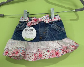 May include: A denim skirt with a floral print ruffle and bow. The skirt has a blue denim base with a pink and white floral print ruffle and bow. The tag reads "BEQUE". The skirt is hanging on a gray hanger.