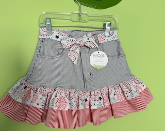 May include: A skirt featuring a grey and white pinstripe pattern with a waistband and ruffled hem decorated in a red, white, and blue star and firework print. The skirt includes a fabric tie at the waist and a circular tag labeled BEQUE.