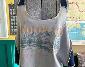 May include: A gray tank top with the word "COLORADO" printed across the front in orange. Below the word is a graphic of mountains and trees. The tank top has dark blue straps and accents.