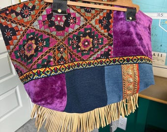 May include: A patchwork tote bag with a wooden handle and leather accents. The bag features embroidered panels in shades of pink, orange, and black, along with sections of purple velvet and denim. The bottom edge is trimmed with long, tan fringe.