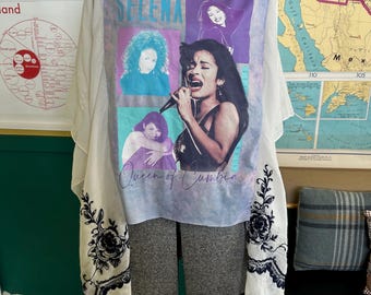 May include: A white t-shirt featuring a graphic of Selena, with the text "SELENA" in blue. The shirt is paired with a sheer, cream-colored overlay with black floral embroidery and tassels. The overlay sleeves are visible on either side of the shirt.