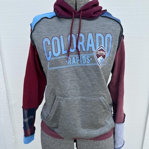 May include: A color-blocked hoodie featuring a heather gray center panel with the text "COLORADO RAPIDS" in light blue and a team logo patch. The garment includes maroon sleeves, a maroon hood with drawstrings, light blue accents, and a plaid cuff.