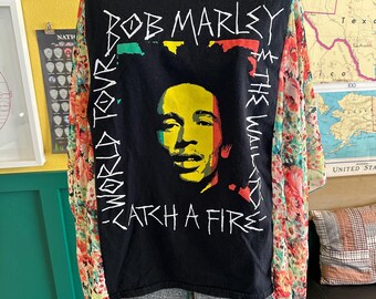 May include: Black t-shirt featuring a Bob Marley portrait with the text "Bob Marley World Tour Catch a Fire". The sleeves are made of a sheer floral fabric in shades of red, orange, and yellow.