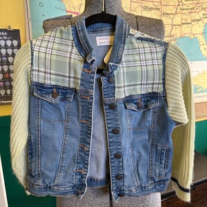 May include: A denim jacket with light blue wash, featuring green and white plaid accents on the shoulders and a light green knit sweater for sleeves. The jacket has button closures and the text "beque textile".