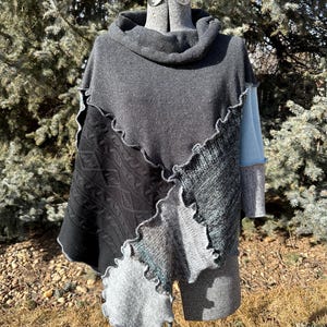 May include: A patchwork poncho in shades of gray, black, and blue, displayed on a mannequin. The poncho features a cowl neck and a mix of textures, including knit and felted sections with a wavy edge. The sleeves are a light blue color.