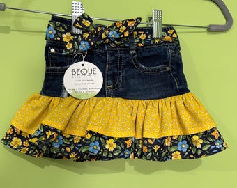 May include: A denim skirt with a yellow floral ruffle and a matching bow. The skirt has a dark blue denim base with a yellow and blue floral pattern on the ruffle and bow. The skirt is hanging on a gray hanger.