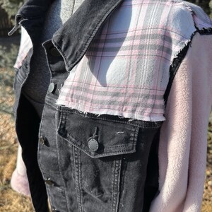 May include: A black denim jacket with a pink and gray plaid pattern on the shoulders and a soft pink fleece sleeve. The jacket has black buttons and a collar.