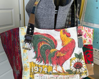 May include: A patchwork tote bag featuring a central panel with a rooster illustration, a 1974 calendar print, and a sunflower. The bag includes side panels of red and floral fabric, a dark brown leather strap with a metal buckle, and decorative stitching.