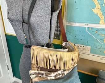 May include: A crossbody bag with a fringed top, brown leather accents, and a blue suede body. The bag features a decorative animal print panel and a long, adjustable strap. The bag is displayed on a mannequin.