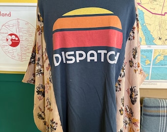 May include: A dark blue t-shirt with the word "DISPATCH" in white, beneath a sunset design in yellow, orange, and red. The sleeves are a light tan fabric with a floral pattern. The shirt is displayed on a mannequin.