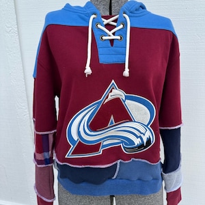 May include: A maroon and blue hooded sweatshirt featuring a large embroidered Colorado Avalanche logo on the front. The garment has a lace-up neckline with white drawstrings and is constructed from various fabric panels including plaid and waffle textures.