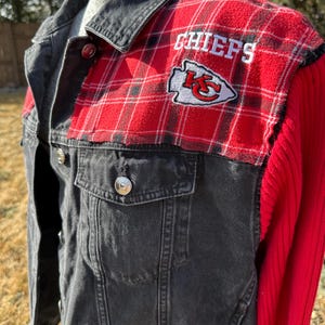 Puede incluir: Chaqueta vaquera negra con detalles de cuadros rojos en los hombros. La chaqueta presenta la palabra "CHIEFS" bordada en blanco, junto con el logotipo del equipo. Las mangas son de punto acanalado rojo liso.