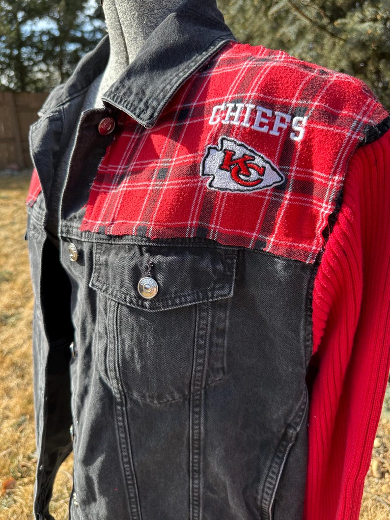Puede incluir: Chaqueta vaquera negra con detalles de cuadros rojos en los hombros. La chaqueta presenta la palabra "CHIEFS" bordada en blanco, junto con el logotipo del equipo. Las mangas son de punto acanalado rojo liso.