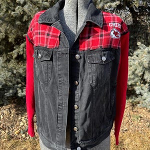 Puede incluir: Chaqueta vaquera negra con detalles de cuadros rojos en los hombros y la parte superior del pecho. Las mangas son de tejido de punto rojo. La chaqueta tiene botones plateados y un parche con la palabra "CHIEFS" y un logotipo.